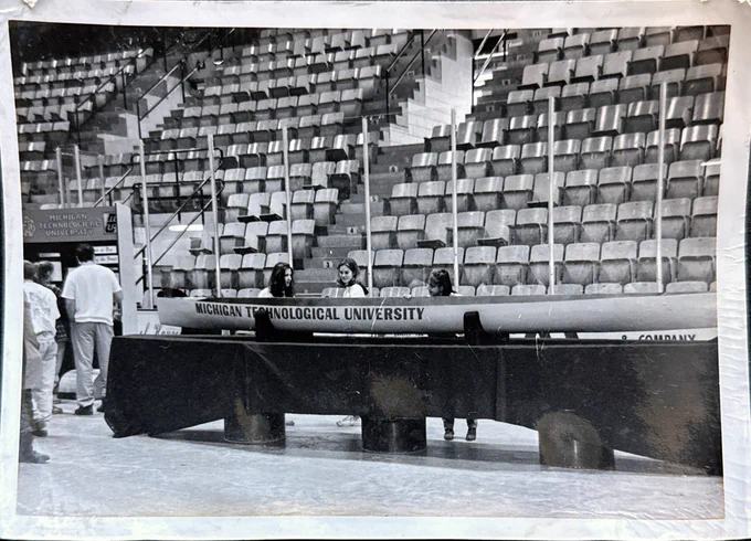 A black-and-white photograph shows a Michigan Technological University concrete canoe displayed on supports inside an arena. Several team members stand alongside the canoe while preparing the display area before competition.
