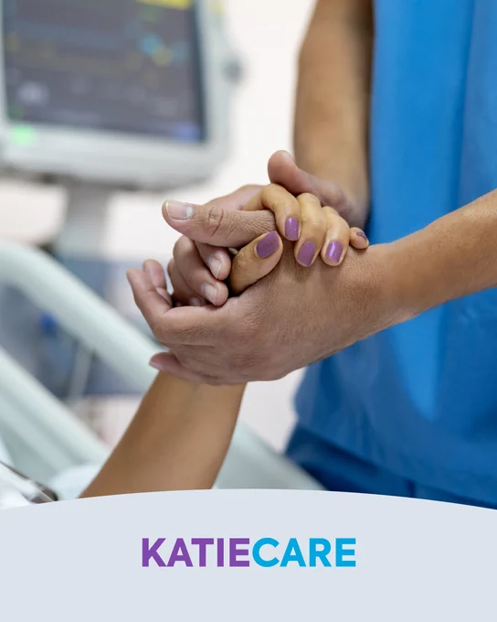 Close-up of a person in blue scrubs holding the hand of another person. A hospital monitor is visible in the background, and a bed in the foreground. Text: KatieCare.