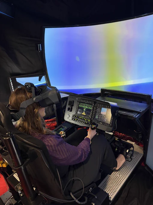 An individual is seated in a simulator cockpit, wearing a virtual reality headset. The curved screen in front displays a colorful gradient. Various controls and monitors are visible inside the cockpit.