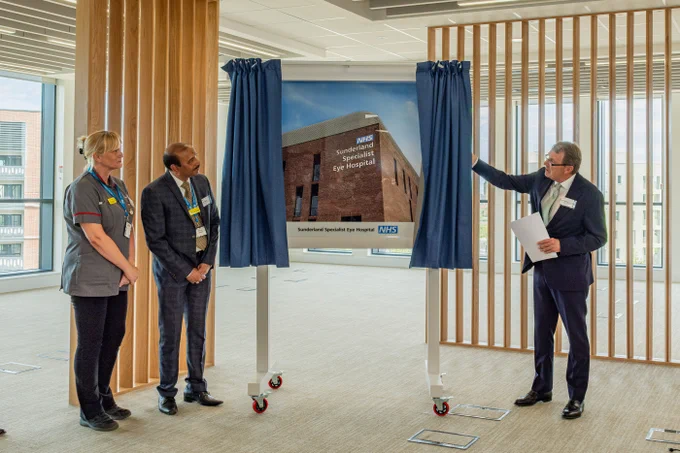 Certainly! Here's an alternative text description:

A group of medical professionals stand in a modern room, unveiling a sign. The sign reads "Sunderland Specialist Eye Hospital" with the NHS logo. The setting is bright, with large windows in the background.