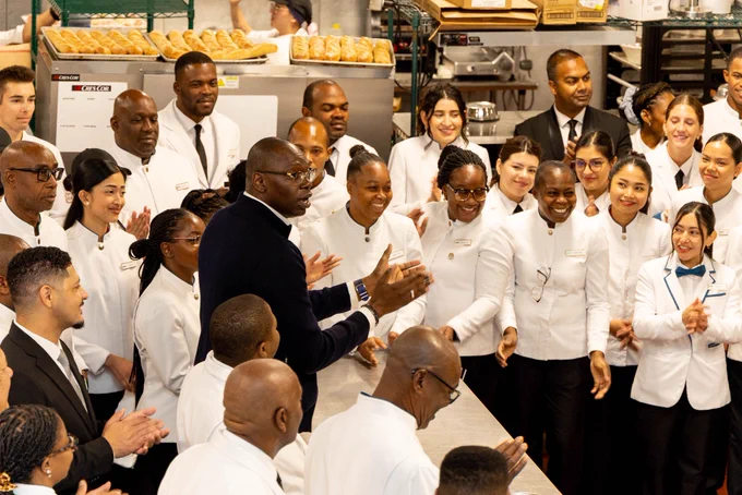 A large group of culinary and hospitality staff wearing white chef coats or uniforms, gathered together in a commercial kitchen setting. In the center, a tall Black man wearing a dark blue jacket and glasses is speaking and gesturing with his hands. The surrounding staff members are smiling, looking toward him, and clapping. Racks of freshly baked bread loaves are visible on equipment in the background.