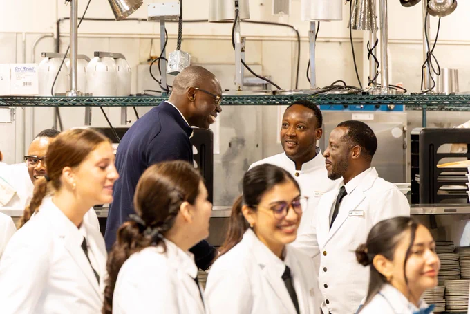 The Lieutenant Governor, wearing a dark navy jacket and glasses is smiling and conversing with two men in white hospitality uniforms within a commercial kitchen environment. In the foreground, several culinary staff members in white coats with black ties are partially visible, smiling and looking off-camera. Metal heating lamps and kitchen equipment are visible in the background.