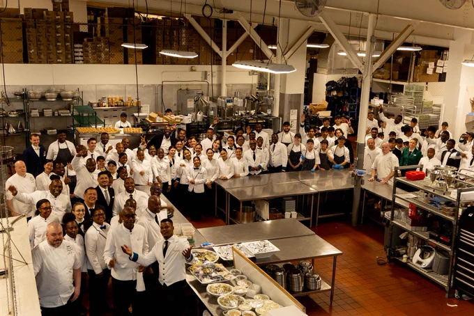 A high-angle, wide shot of a massive team of kitchen staff, chefs, and other staff posing together for a group photo inside a large commercial kitchen. Most individuals are wearing white chef uniforms, and they are arranged around stainless steel prep tables. In the lower left foreground, a man in a white jacket gestures welcomingly toward food prep stations, while the rest of the large crowd fills the spacious, industrial kitchen area extending into the background.