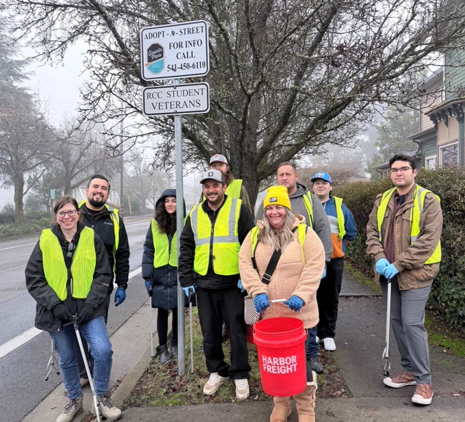 A group of RCC student veterans and employes wearing yellow safety vests and gloves are standing on a sidewalk next to a road doing street cleanup. One person is holding a red Harbor Freight bucket. They are gathered near a sign that reads "Adopt-A-Street: RCC Student Veterans." It's overcast with bare trees in the background.
