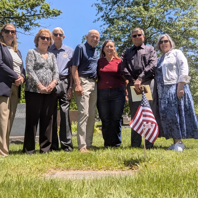 Group of seven adults standing together outdoors on a sunny day near a gravestone with an American flag planted in the grass.