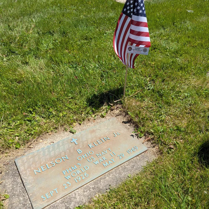 Flat grave marker for Nelson B. Klein Jr., a World War II US Navy veteran from Ohio, with an American flag planted nearby in green grass.