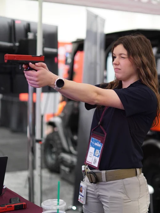 A person at an indoor exhibition booth is holding a training gun and aiming at a screen. They are wearing a smartwatch and a badge on a lanyard. In the background, there is a Yamaha vehicle and TEEX employee watching behind her.