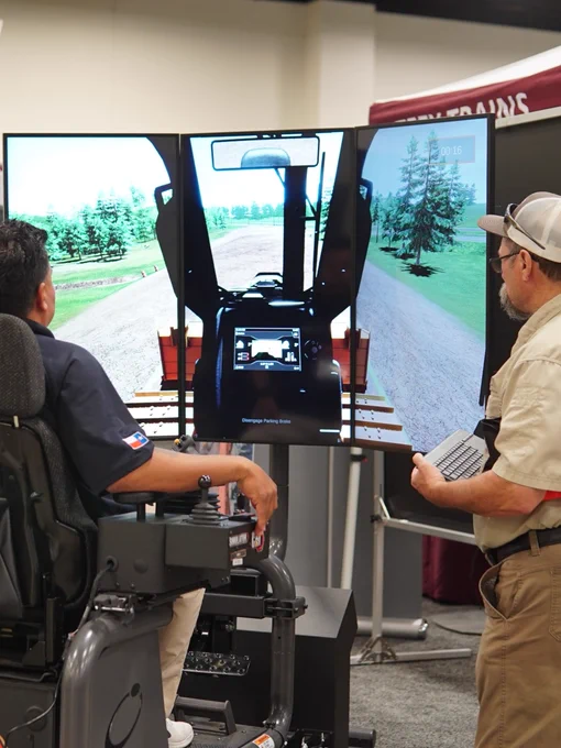 A person is seated in a simulator chair branded with the CAT logo, engaging with a large screen displaying a virtual construction environment. Two individuals stand nearby, observing the setup. The scene is set in an indoor exhibition space, with informational displays and banners in the background.
