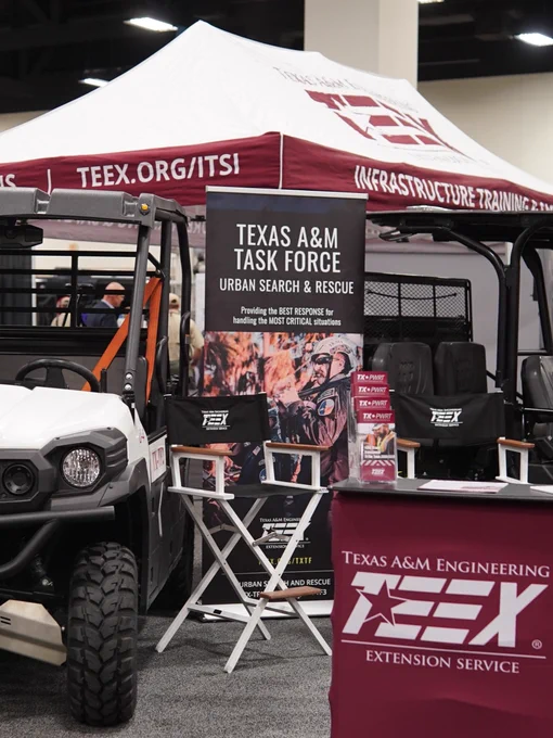 A trade show booth featuring an off-road vehicle with bars and the logo "TEEX." The booth is labeled "Texas A&M Task Force" and "Texas A&M Engineering Extension Service," showcasing urban search and rescue information. Nearby are brochures and a display area.