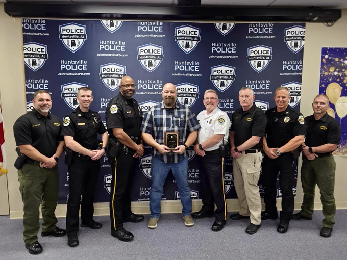 A group of men in police uniforms and civilian clothing stand in a line in front of a Huntsville Police Department backdrop. The person in the center is holding a plaque, and the others are standing on either side in a pose for a formal recognition photo. The backdrop displays multiple Huntsville Police Department logos and website text.