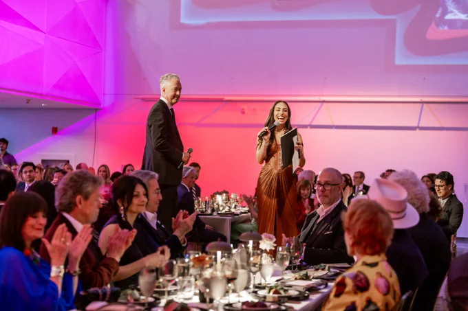 Two people standing in the middle of a room with many seated people and ambient lighting. The man on the left wearing a black suit, the woman on the right wearing a bronze dress and speaking into a microphone. 
