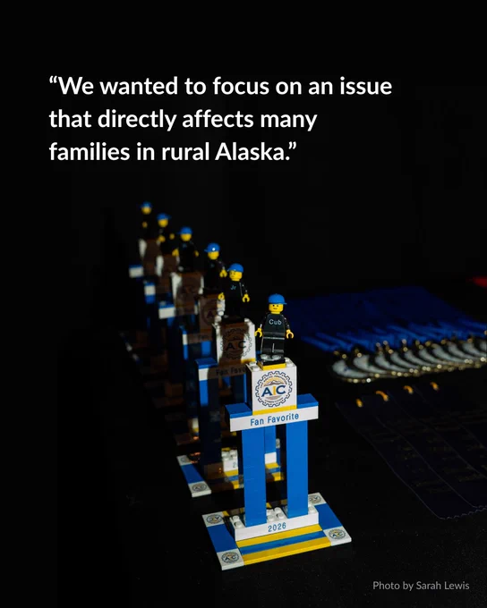 Blue and yellow trophies resembling LEGO bricks stand in a row, each topped with a small toy figure. The front trophy is labeled "Fan Favorite 2026" with a logo resembling a gear. To the right, blue ribbons are laid on a table. Overhead, white text reads, “We wanted to focus on an issue that directly affects many families in rural Alaska.” Photo credit: Sarah Lewis.