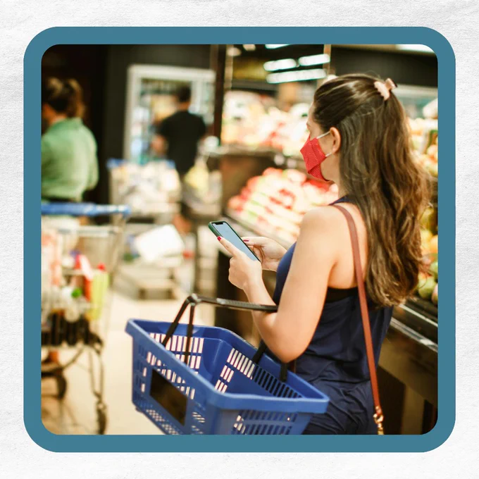 A woman with a red mask holds a smartphone and a blue shopping basket in a grocery store.