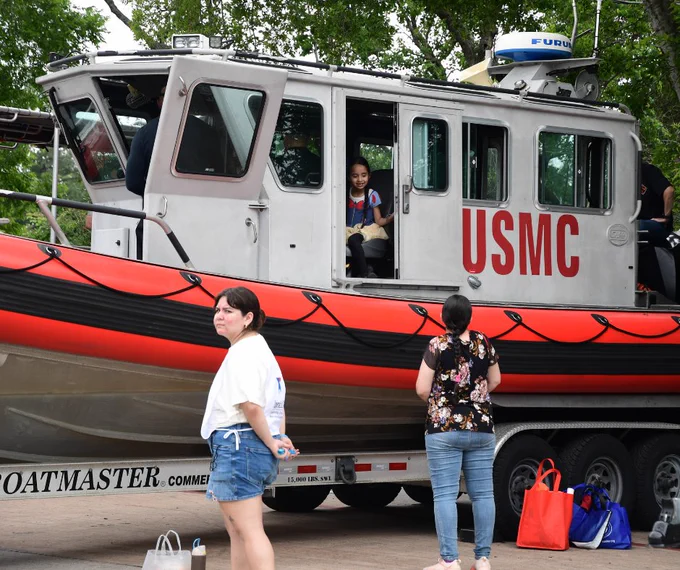 A person stands near a USMC boat on a trailer. Another individual is inside the boat's cabin. Two others are nearby, with various bags on the ground. Trees are in the background.