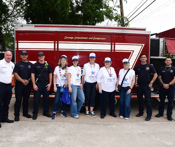 A group of people, including uniformed firefighters and individuals wearing various Love to the Rescue shirts, stand in front of a fire truck. Some are wearing hats, and one person is holding a small dog.