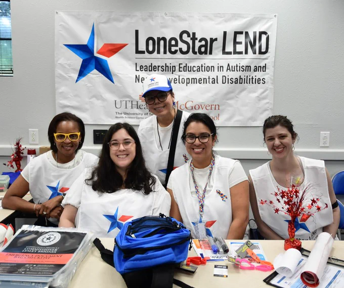 Five individuals are sitting at a table, smiling in front of a "LoneStar LEND" banner, which stands for Leadership Education in Autism and Neurodevelopmental Disabilities. The table has various items, including a blue backpack. They appear to be in an educational or organizational setting at UTHealth McGovern Medical School.