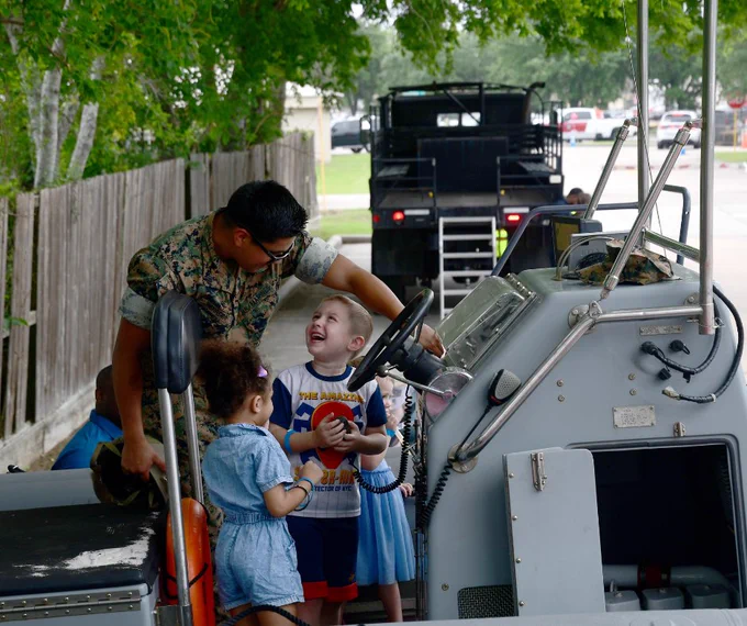 A person in a military uniform interacts with three children on a small boat. One child is holding the wheel, smiling up at the person. A large military vehicle is visible in the background. Trees and a fence line the setting.