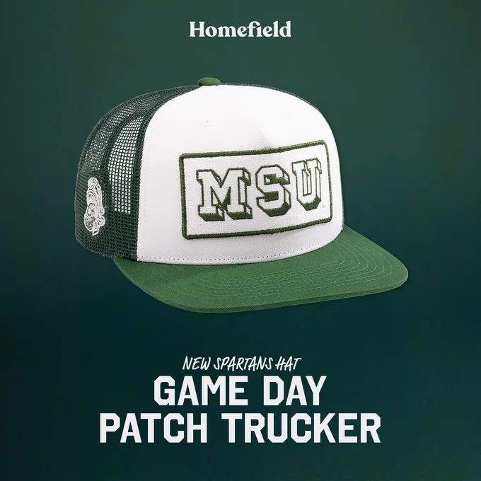 A trucker hat with a green visor and mesh back features "MSU" on the front and Gruff Sparty image on the side. The Homefield logo is at the top, and the phrase "New Spartans Hat Game Day Patch Trucker" is displayed below the hat.