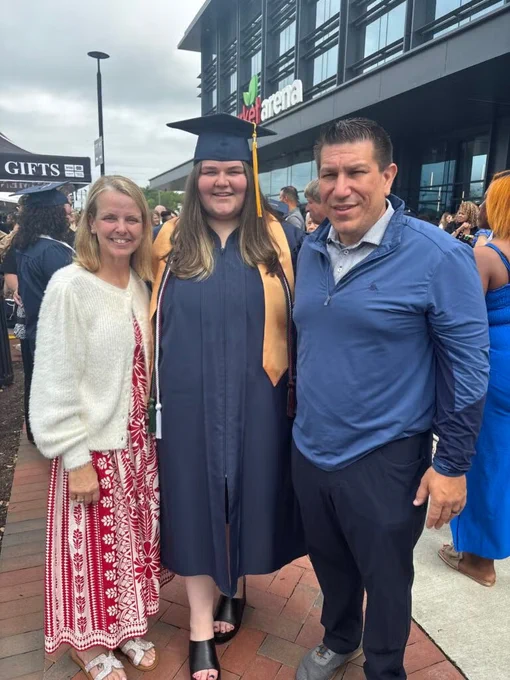 A person in a graduation cap and gown stands smiling between two others outside a building. A crowd is in the background.