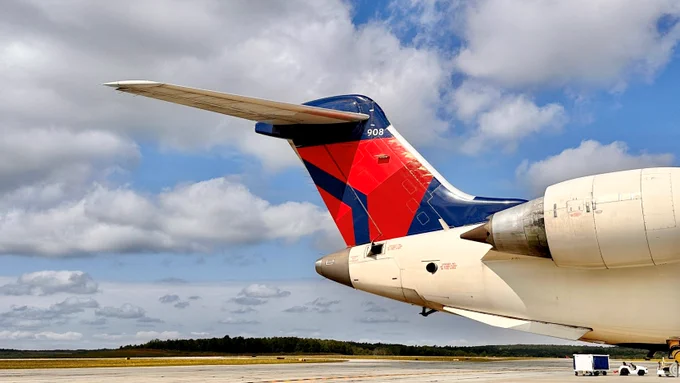 The image shows the tail section of a Delta Air Lines aircraft on an airport tarmac. The tail displays Delta's logo in blue and red. A small vehicle is visible in the background under a partly cloudy sky.