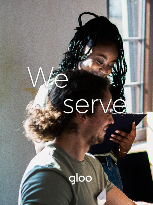 Two people in an office setting collaborate, with text 'We Serve' overlaid. The scene is warm and friendly.
