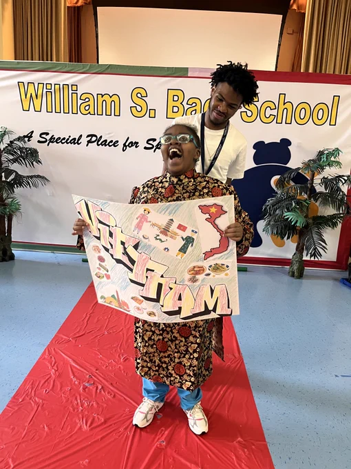 Student standing on a red carpet holding a decorated poster in front of a William S. Bach School banner with plants on either side.