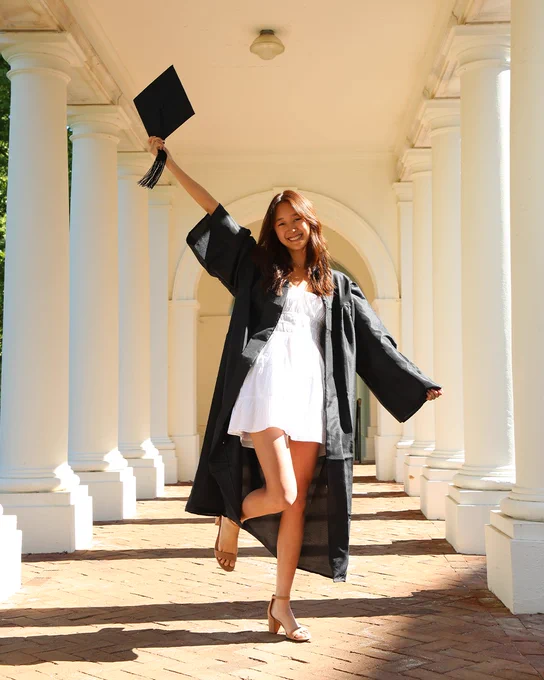 Kaylee, smiling and jumping in the air while wearing a graduation cap and gown and white dress.
