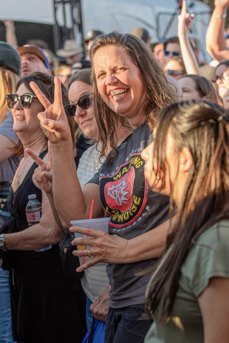 A woman in a crowd smiles and gives the peace sign.