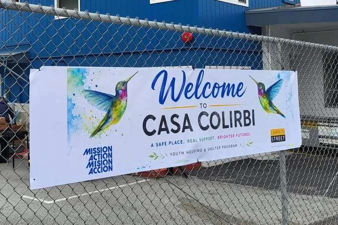 Welcome sign for Casa Colibrí displayed on a fence outside the housing site, featuring hummingbird artwork and Mission Action and Larkin Street Youth Services logos.