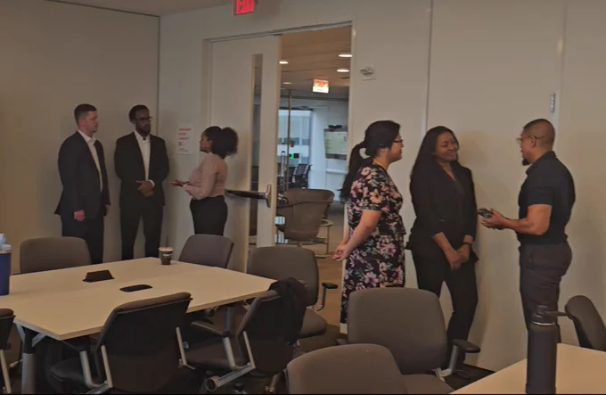 Six professionals engaged in small group conversations inside a modern office meeting room with tables and chairs.