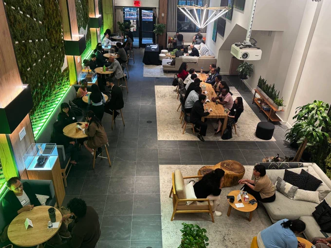 Modern coworking space with people working and chatting at tables and sofas, featuring green plant walls and warm lighting.