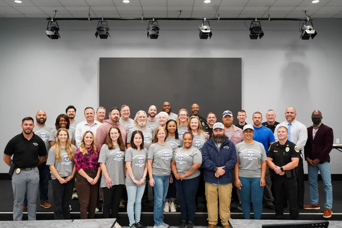 A large group of people is posed for a group photo inside a room with gray walls and overhead studio lighting. Several individuals in the front row wear matching gray shirts with a printed emblem, while others in the group are dressed in assorted professional and casual clothing. The group stands on risers in front of a blank dark panel on the wall.