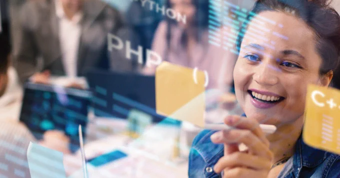 A person smiles while interacting with a translucent digital screen displaying programming languages such as Python, C  , and PHP. Other individuals are visible in the background, blurred.