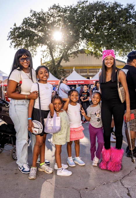 A group of women and girls smile in front of a crowded vendor marketplace.