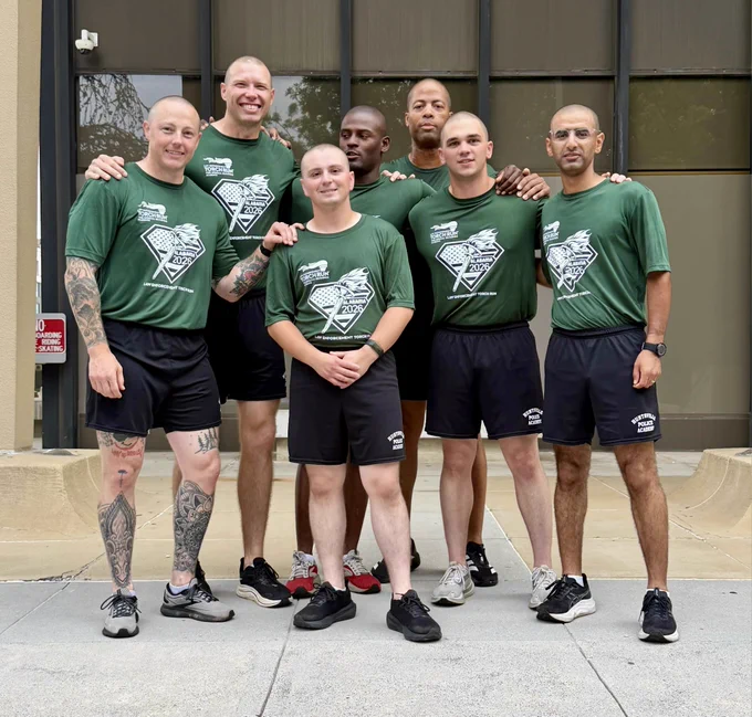 A group of seven individuals is standing together outdoors in front of a building with large windows. All are wearing matching green athletic shirts with a printed logo and black shorts. The group is posed closely, with some individuals standing and others slightly leaning toward the center, and the setting appears to be on a sidewalk or concrete area.