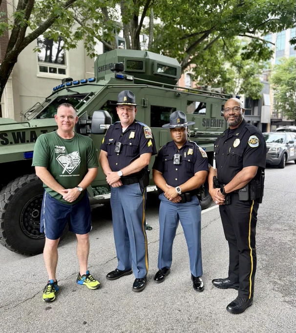 Four individuals are standing on a city street in front of a large green armored S.W.A.T. vehicle. Three of them are wearing dark uniforms with badges and duty belts, while the fourth is wearing athletic clothing and sneakers. Trees, parked cars, and buildings line the street in the background.