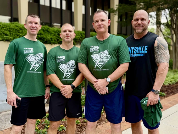Four individuals are standing together outdoors in front of a building with large windows. Three of them are wearing matching green athletic shirts with a printed logo and black shorts, while the fourth is wearing a black sleeveless shirt with blue lettering and holding a green item in one hand. The group is posed on a sidewalk with greenery and trees in the background.