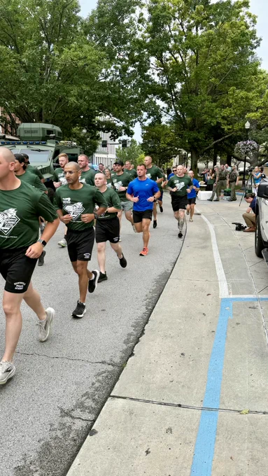 A large group of runners is moving down a city street during an organized run. Most participants are wearing matching green athletic shirts, while a few are in blue shirts. Trees, parked vehicles, and bystanders line the sidewalk, and the atmosphere appears energetic and active as the group moves forward.