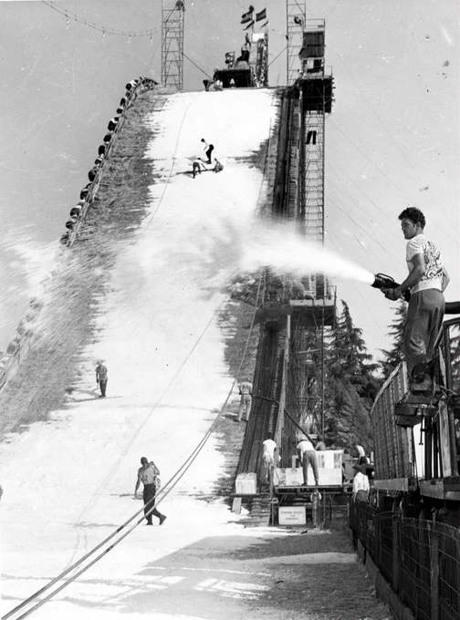 Black-and-white photograph of a massive artificial ski jump at the Los Angeles County Fair in 1951. Workers spray water and ice onto the steep ramp to create snow, while several people stand on the slope and around the structure. The towering jump, supported by scaffolding and lift machinery, rises high above the fairgrounds, illustrating the scale of the temporary attraction built from millions of pounds of ice.