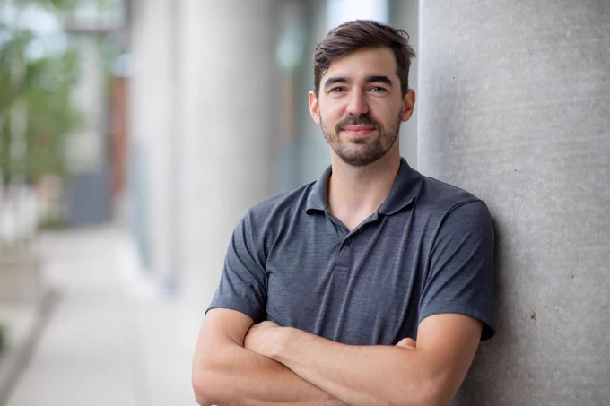 Professor Luka Milosevic stands with arms crossed, leaning against a concrete column outside a building. 