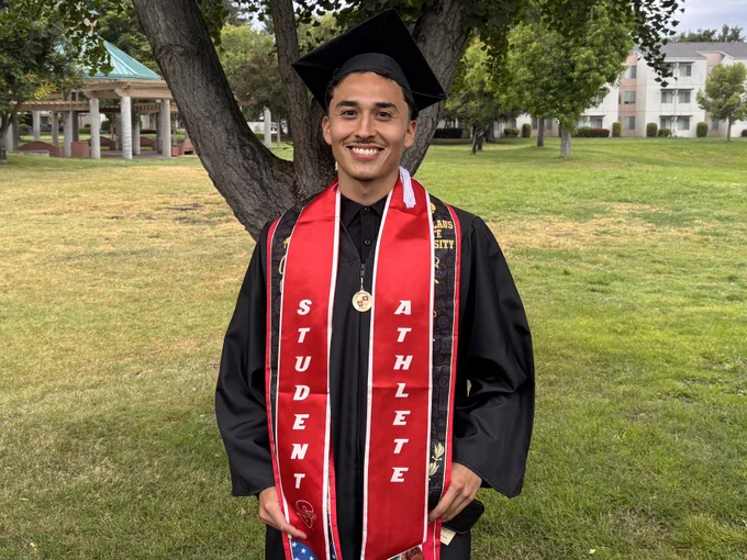 A Stan State graduate poses outdoors wearing a cap and gown, honor cords, a graduation medal, and a red student-athlete stole.
