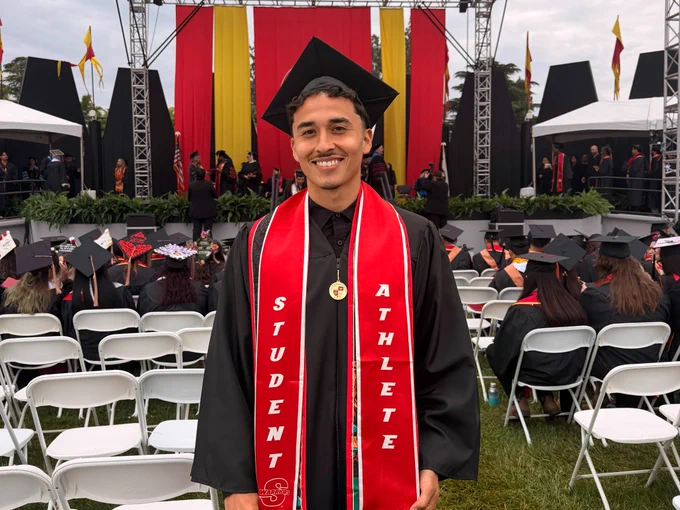 A Stan State graduate poses outdoors beneath a tree wearing a cap and gown, graduation medal, and red student-athlete stole.
