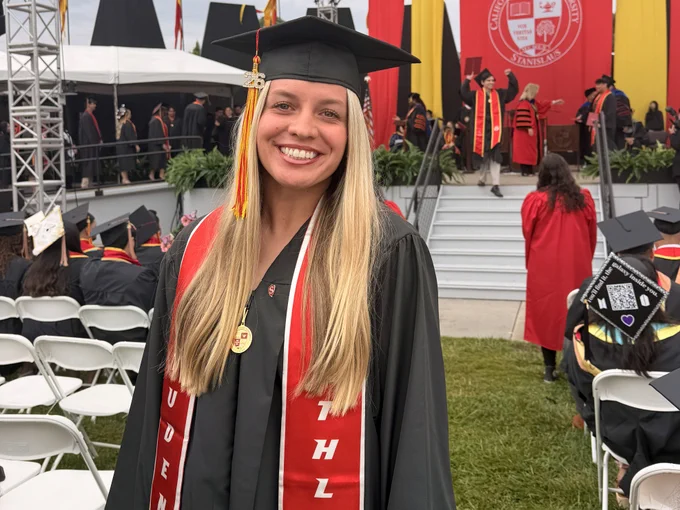 A Stan State graduate smiles in front of the commencement stage wearing a cap and gown, graduation medal, and red student-athlete stole.