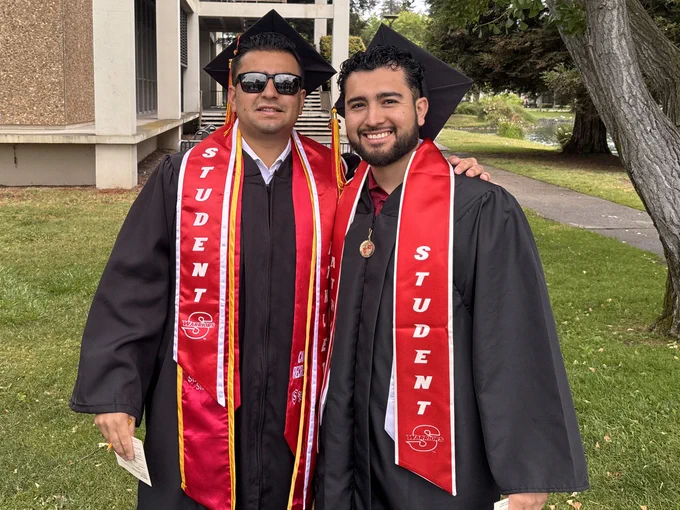 Two Stan State graduates stand together outdoors in caps and gowns wearing red student-athlete stoles and graduation medals.