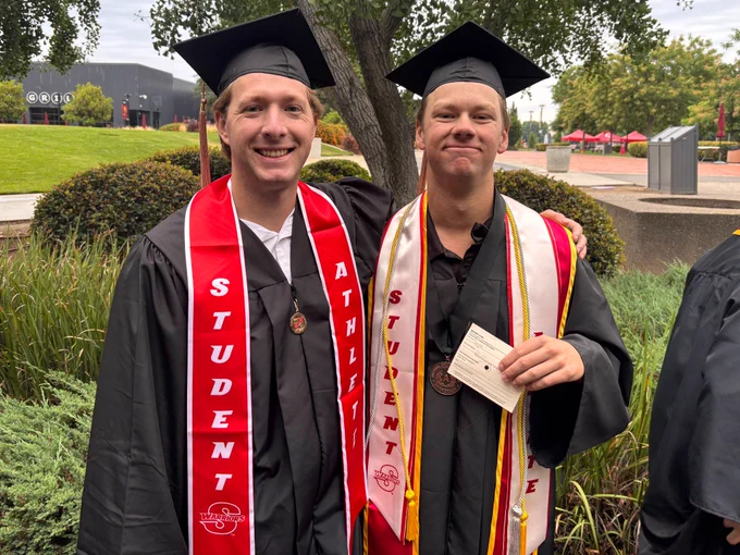 Two Stan State graduates pose together outdoors wearing caps, gowns, medals, honor cords, and red student-athlete stoles.