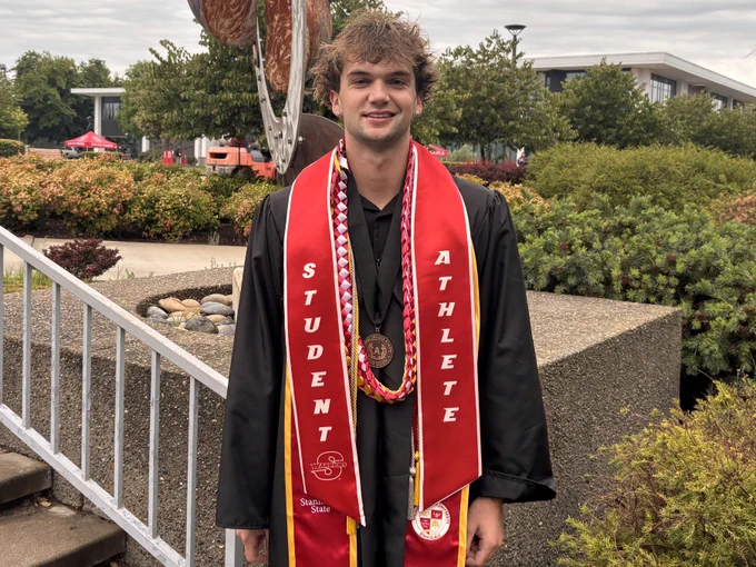 A Stan State graduate poses outdoors wearing a cap and gown, honor cords, a graduation medal, and a red student-athlete stole.