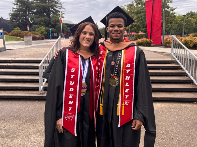Two Stan State graduates stand together outdoors wearing caps and gowns, CCAA championship medals, honor cords, and red student-athlete stoles.