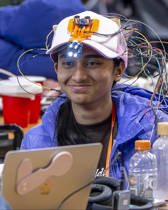 A StarkHacks participant smiles at a hackathon table wearing a baseball cap covered in circuit boards, sensors, and a tangle of colorful wires, with a laptop and Gatorade bottle in the foreground.