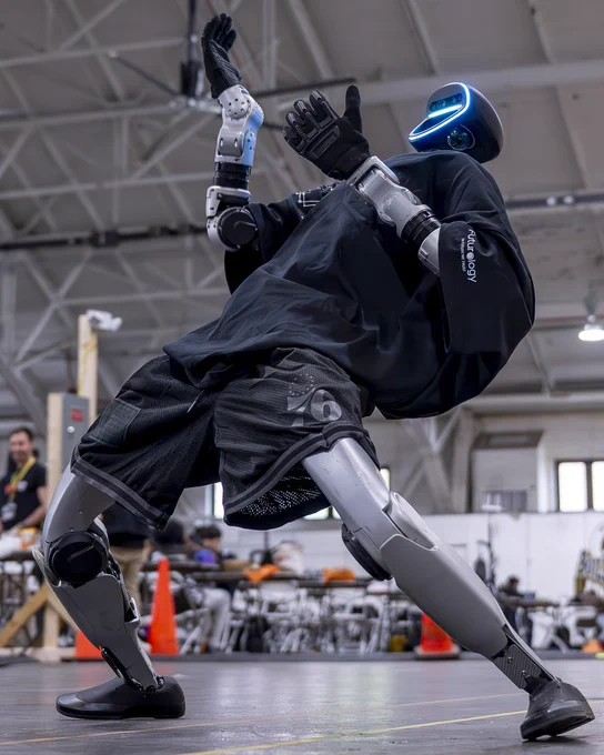 A humanoid robot wearing a black cape and shorts strikes a dynamic wide-stance pose on the floor of the Purdue University Armory during StarkHacks, with hackathon participants working at tables in the background.