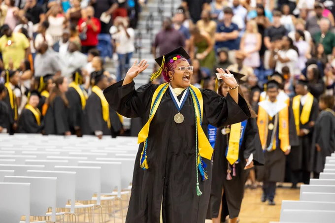 A smiling graduate in a black cap and gown, holding a diploma, stands among a crowd of other graduates at a commencement ceremony.